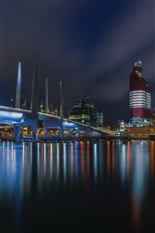 Byens natlige skønhed - Oplev den fortryllende atmosfære fra en by oplyst af natten, hvor moderne arkitektur reflekteres i det stille vand. Denne levende scene skaber en unik dybde og elegance i dit hjem.
